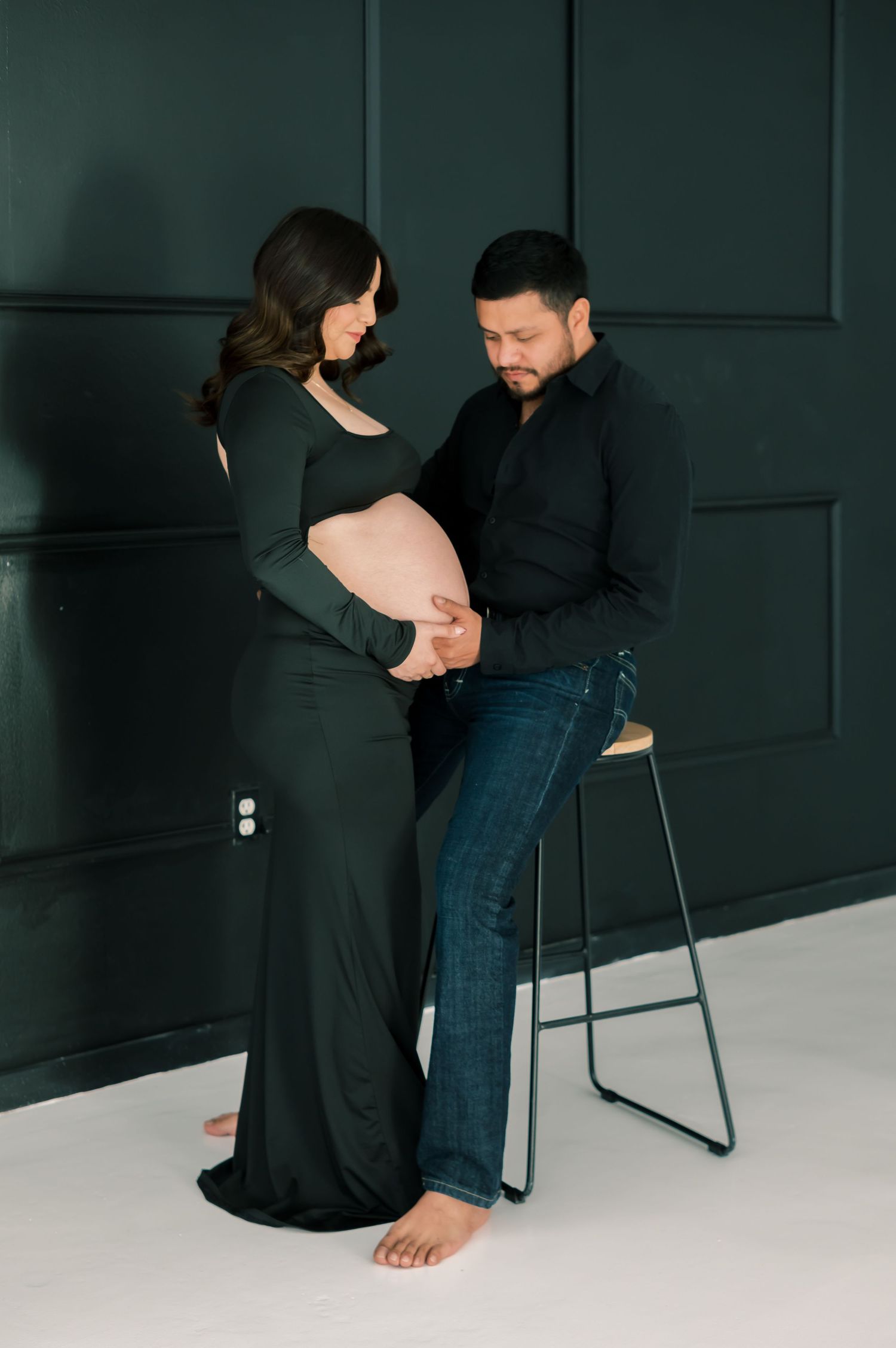 Expecting couple in matching black outfits pose together in studio, man seated on stool tenderly touching pregnant woman's belly.