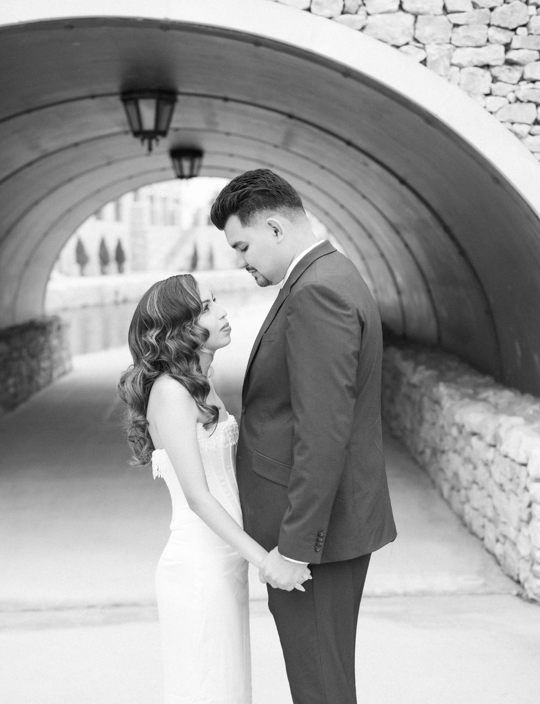 Black and white romantic engagement photo of couple holding hands under stone arch tunnel.