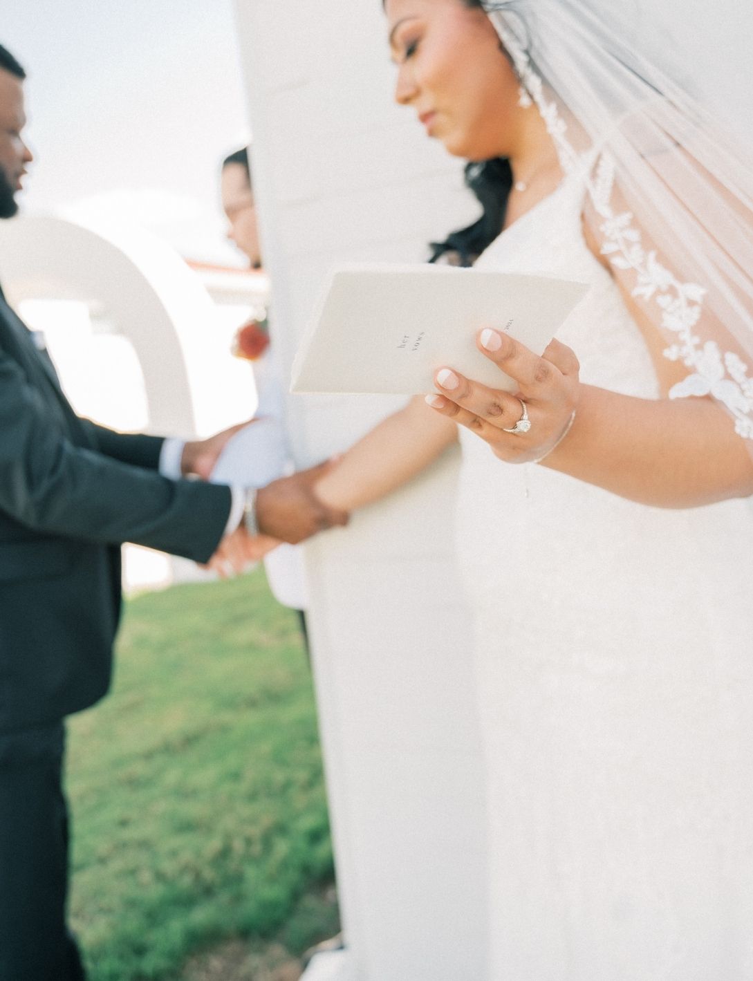 Bride in lace wedding gown and veil reading vows from a card while holding grooms hand at outdoor ceremony.