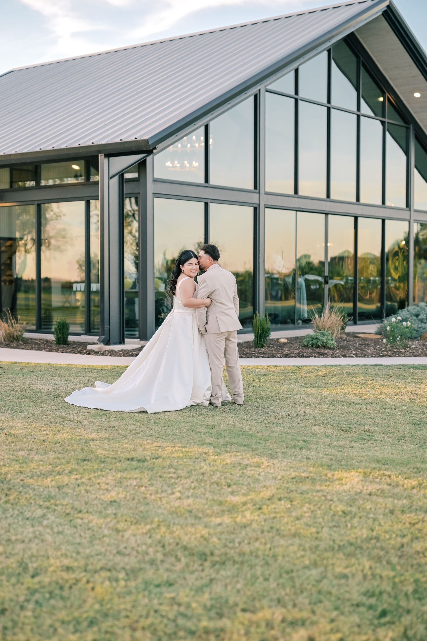 Modern wedding venue with glass walls and metal roof surrounded by manicured lawn.