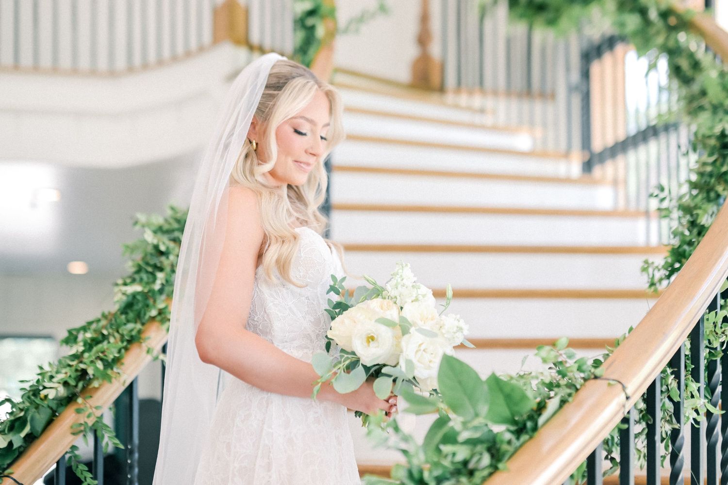 Bride in lace gown holds white bouquet on a staircase decorated with lush greenery garland.