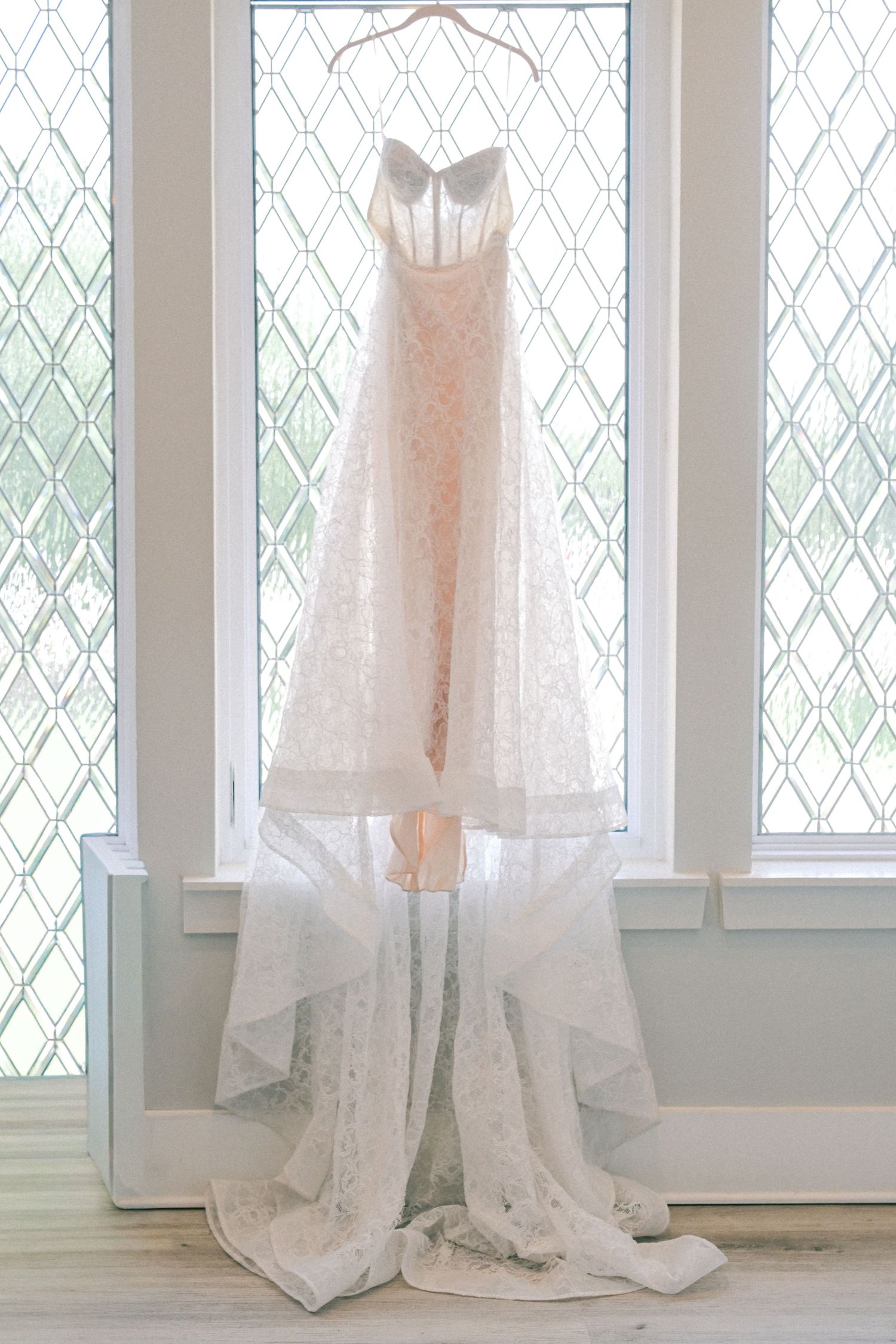 Elegant lace wedding dress hanging in front of leaded glass windows with natural light.