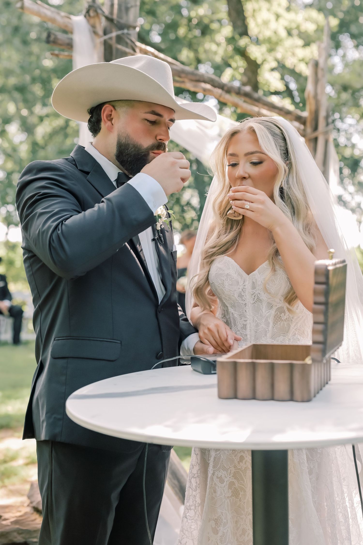 Bride and groom in cowboy hat share communion during outdoor wedding ceremony at rustic venue.