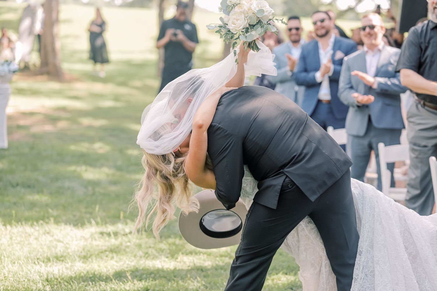Groom dips bride for a romantic kiss outdoors as wedding guests cheer and applaud.
