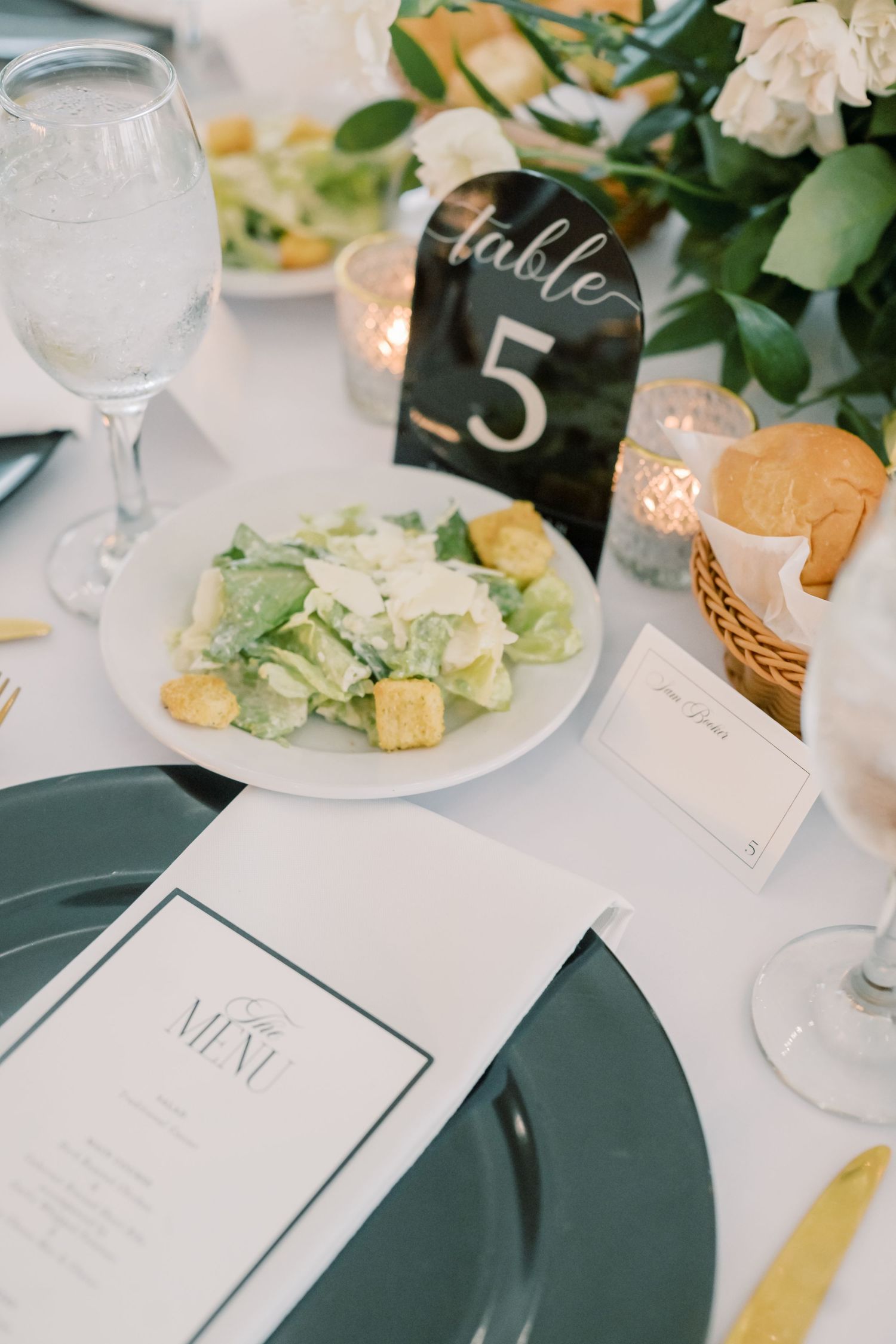 Elegant wedding reception table setting with Caesar salad, green charger plates, Table 5 sign, and white floral centerpiece.