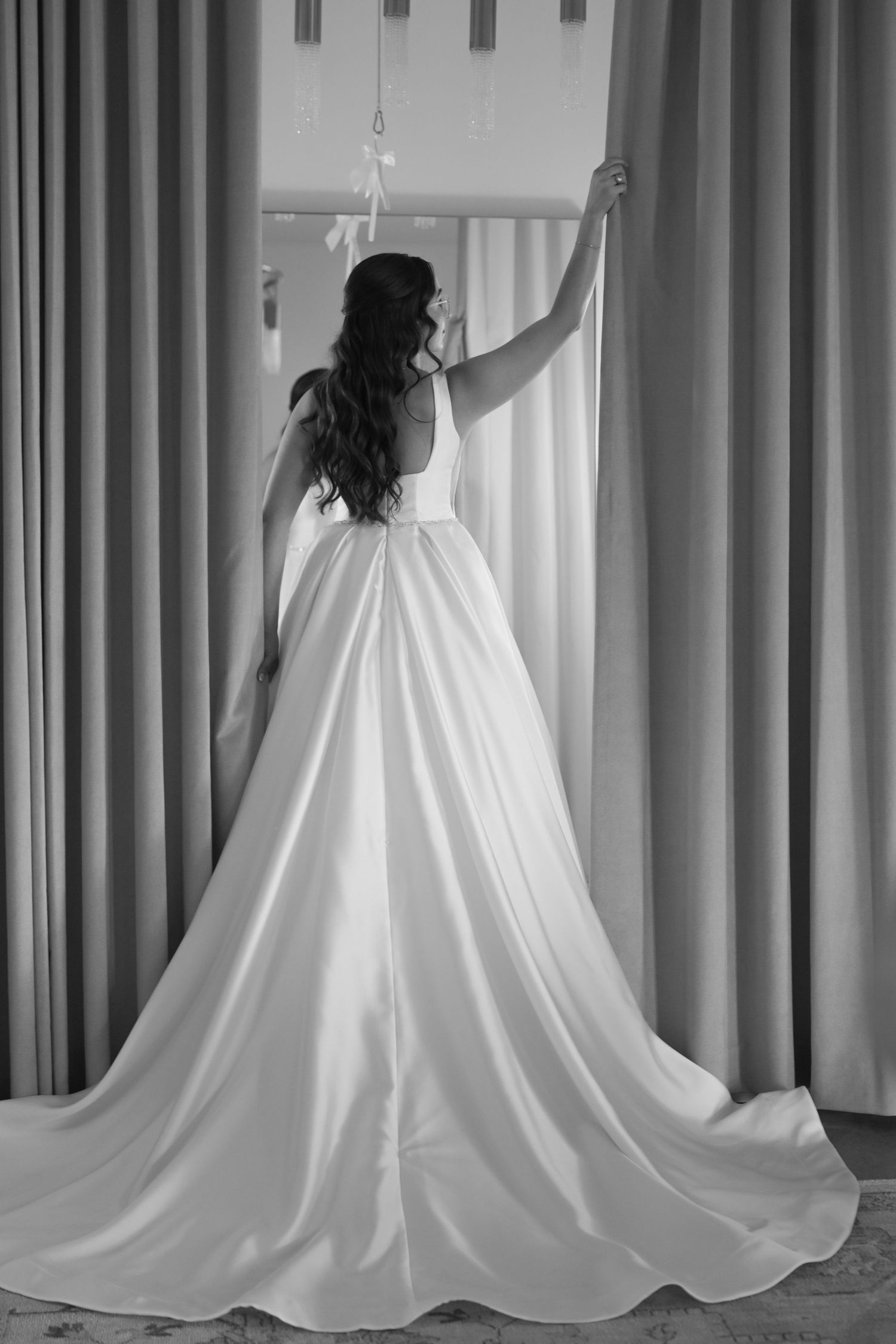 Bride in elegant white satin ball gown with long train standing by curtains from behind.