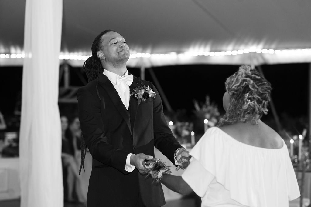 Groom in tuxedo laughing joyfully while holding hands with bride during wedding reception under tent.