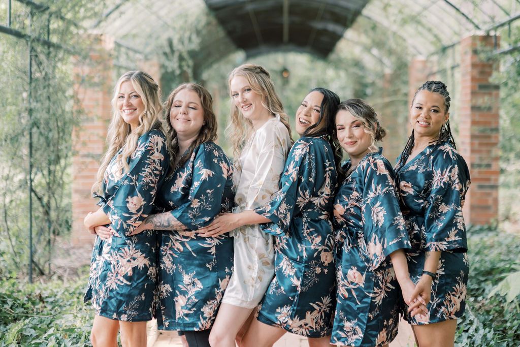 Bride in white robe with bridesmaids in navy floral robes posing together in a lush greenhouse setting.
