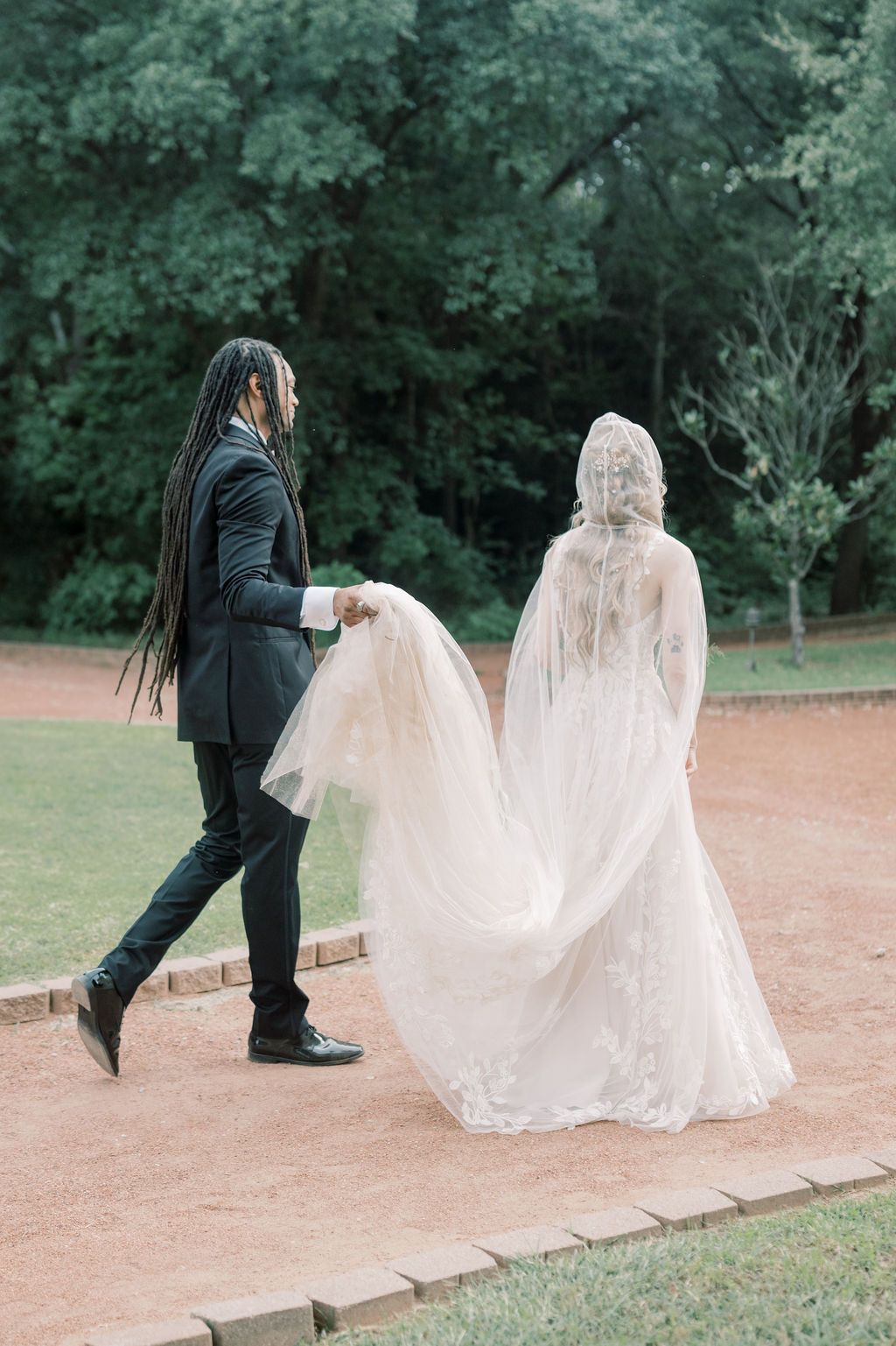A groom with long dreadlocks in a black suit carries his bride's flowing white lace wedding dress train outdoors.