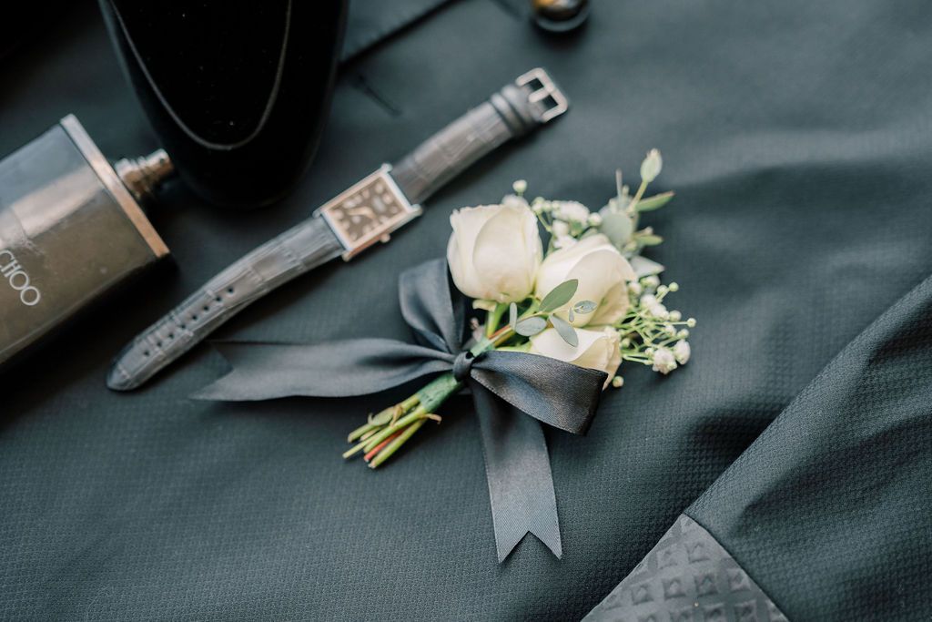 Elegant white rose boutonniere with greenery and dark ribbon beside a watch and cologne on gray suit.