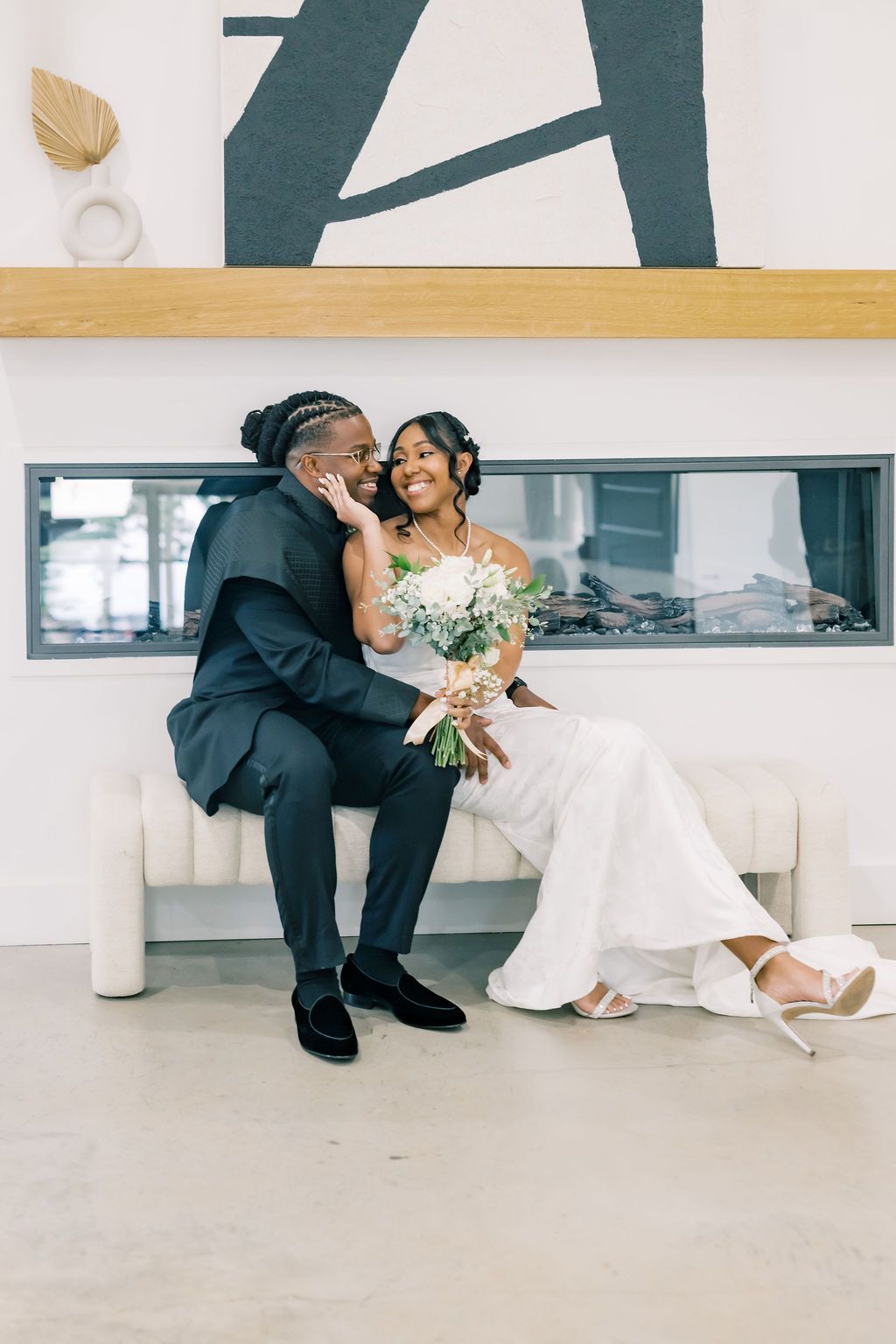 A joyful Black couple in wedding attire sit together on a white bench, the groom whispering to the smiling bride holding a bouquet.