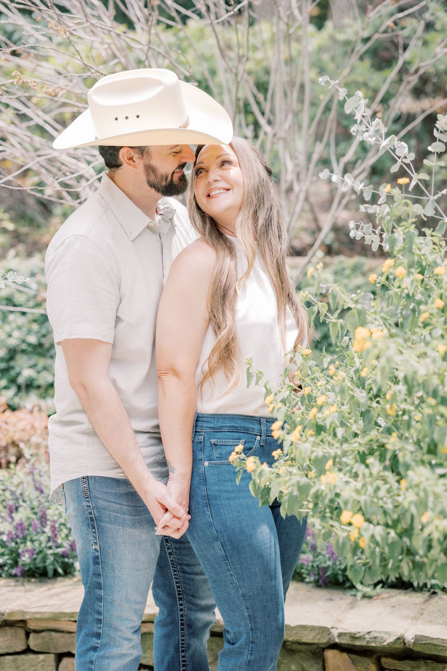 Romantic couple holding hands outdoors, man in white cowboy hat kissing woman's cheek near flowering bushes.