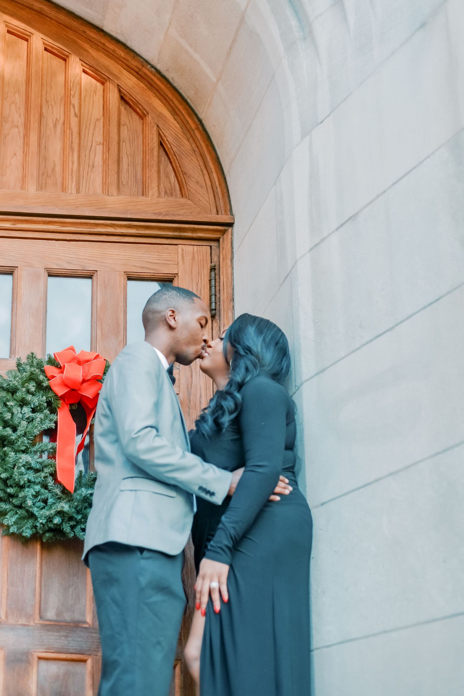 Couple sharing a romantic kiss in front of a wooden arched door with a Christmas wreath decoration.