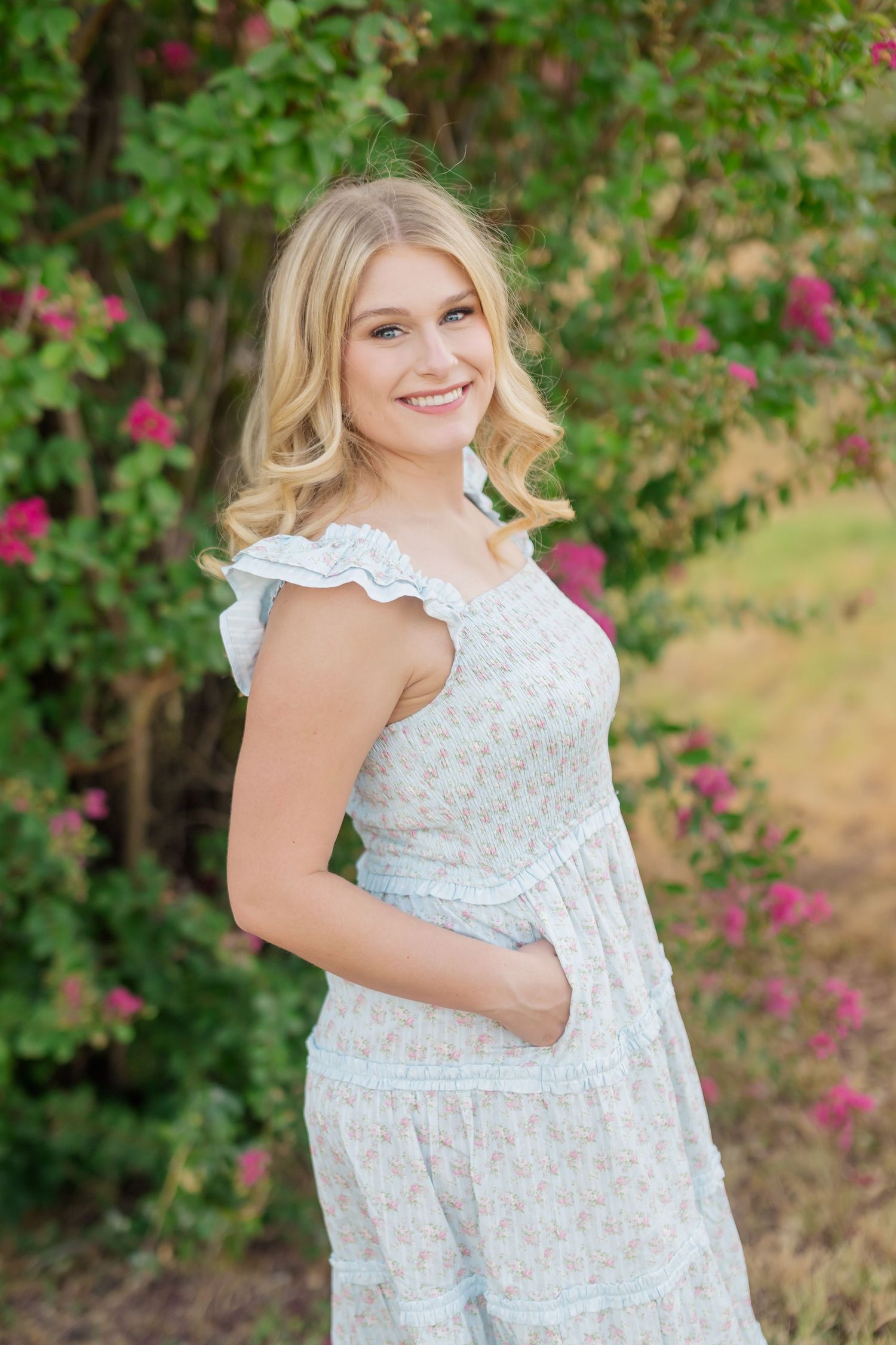 Smiling blonde woman in a floral sundress poses outdoors among blooming pink flowers and lush green foliage.