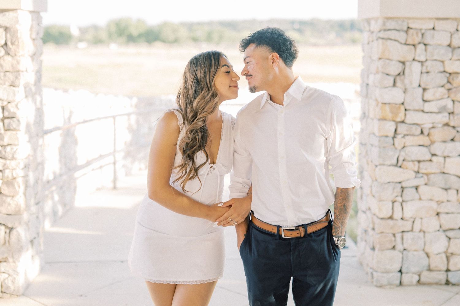 A couple in white outfits share a romantic moment, smiling at each other near stone pillars outdoors.