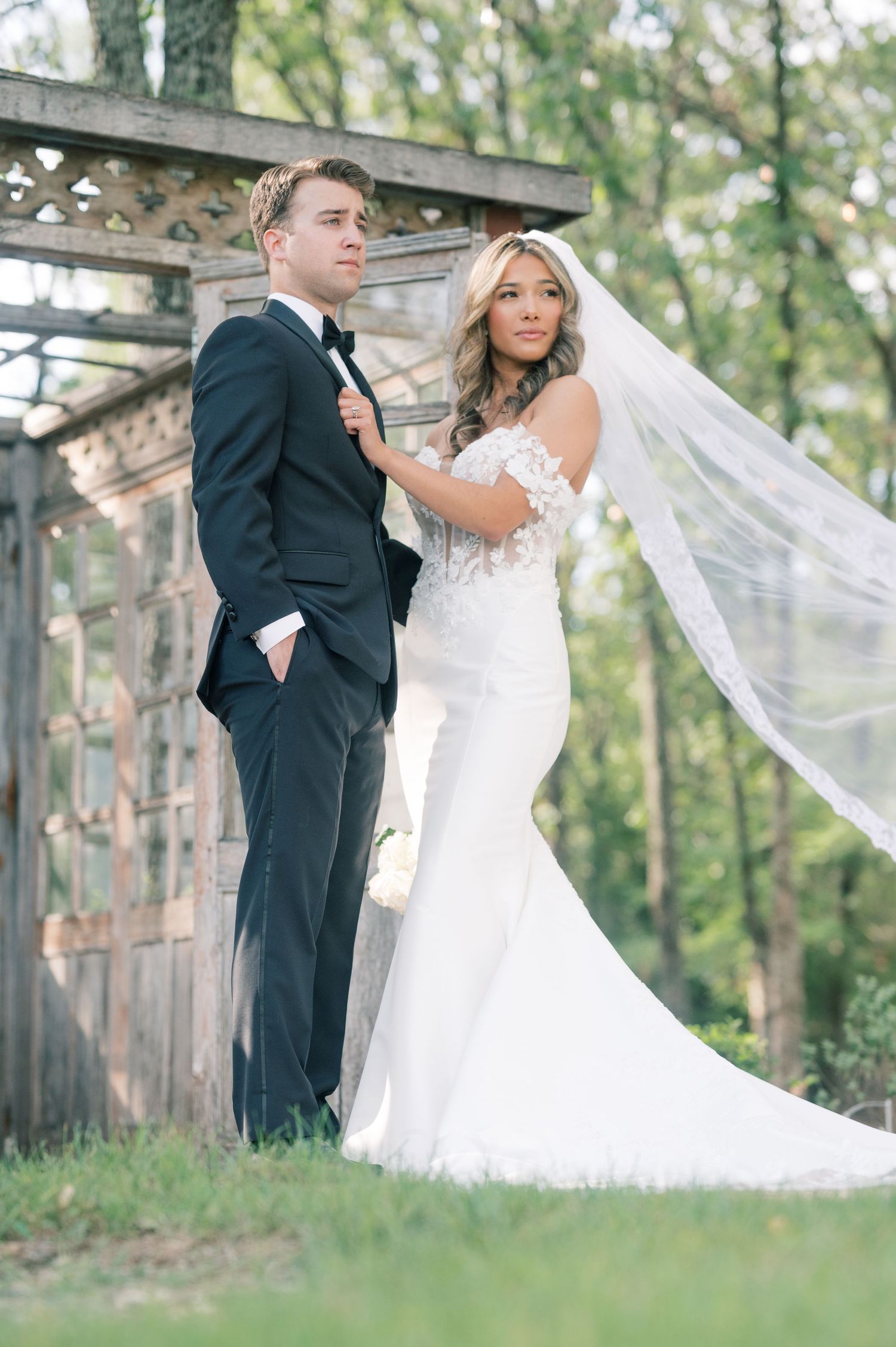 Bride in lace gown and veil stands with groom in black tuxedo outdoors near rustic wooden structure.