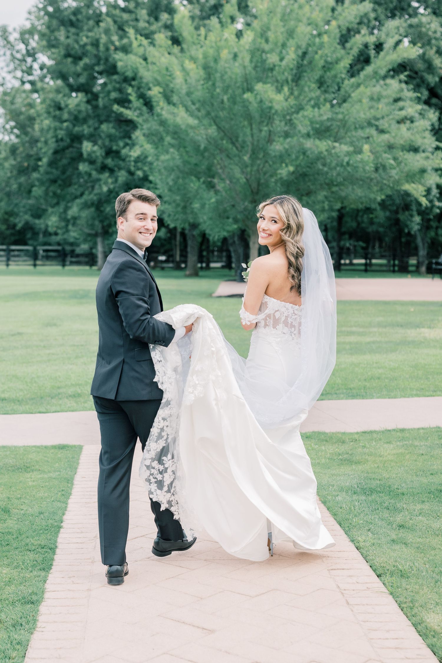 Bride and groom walking on a garden path, groom holding bride's lace wedding veil train outdoors.