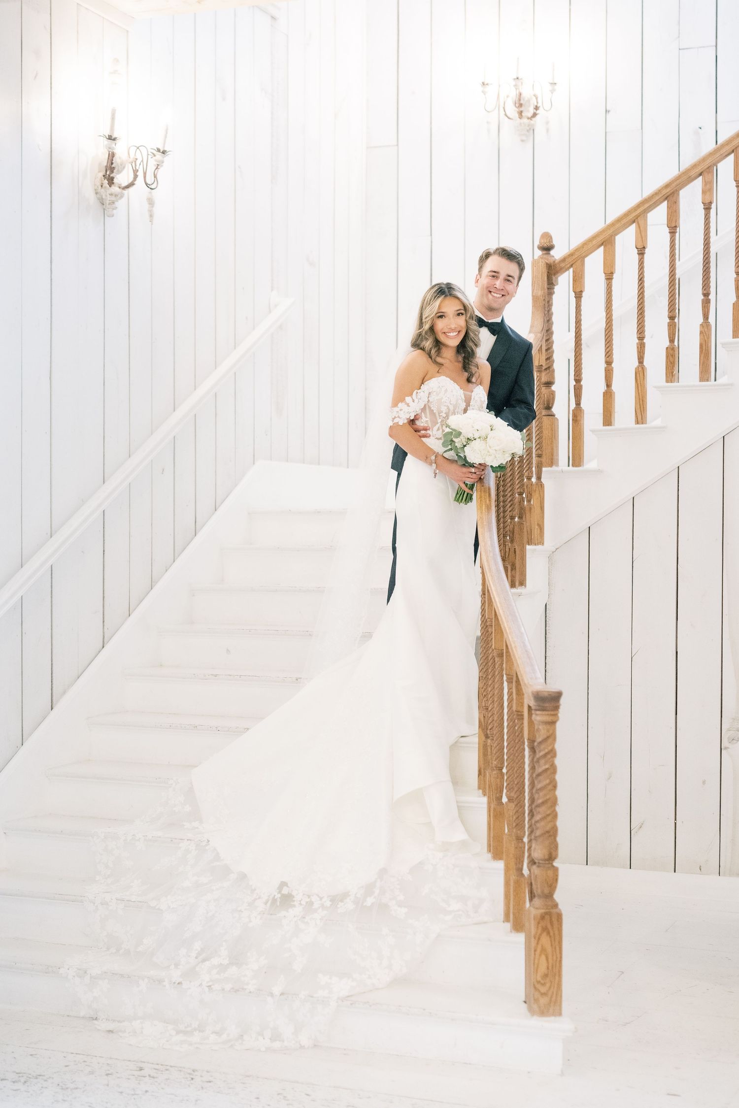 Bride in lace mermaid gown and groom in black tuxedo posing together on a white staircase.