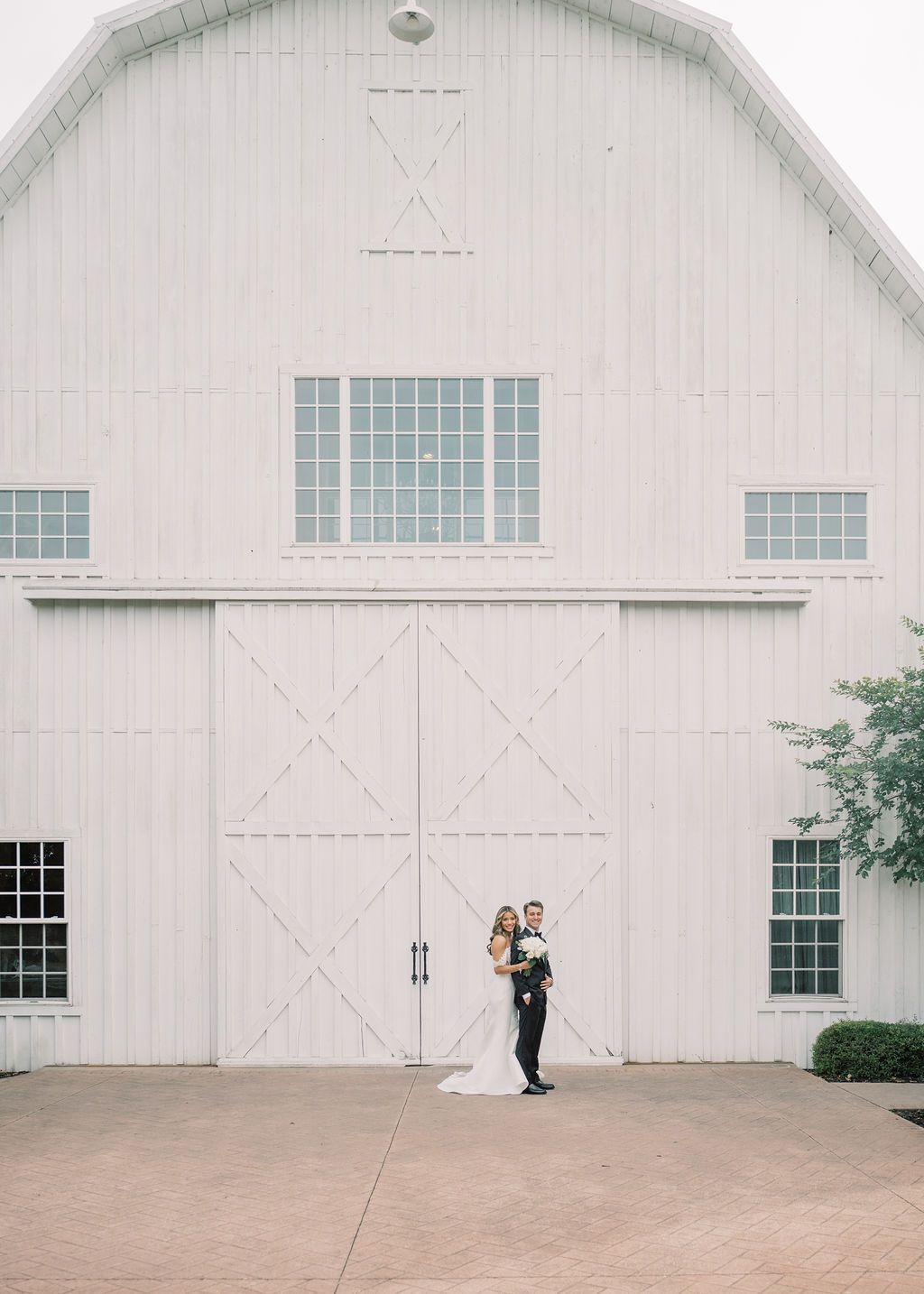 Wedding couple poses before large white barn venue doors on their special day.
