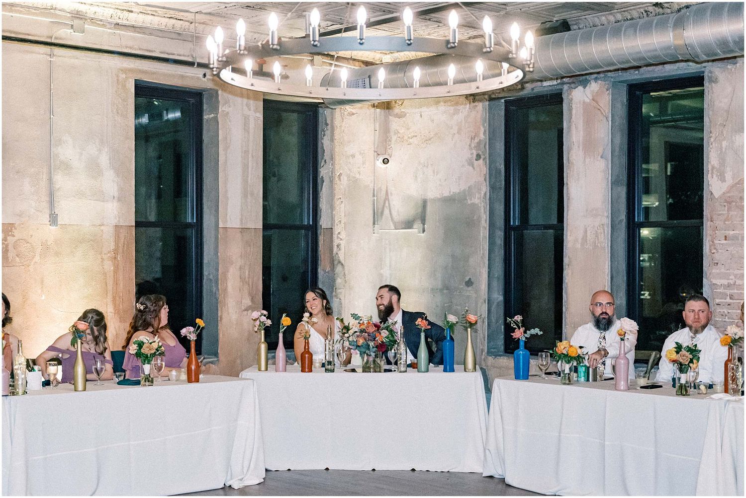 Wedding reception venue with long tables and elegant chandelier lighting in an industrial space.