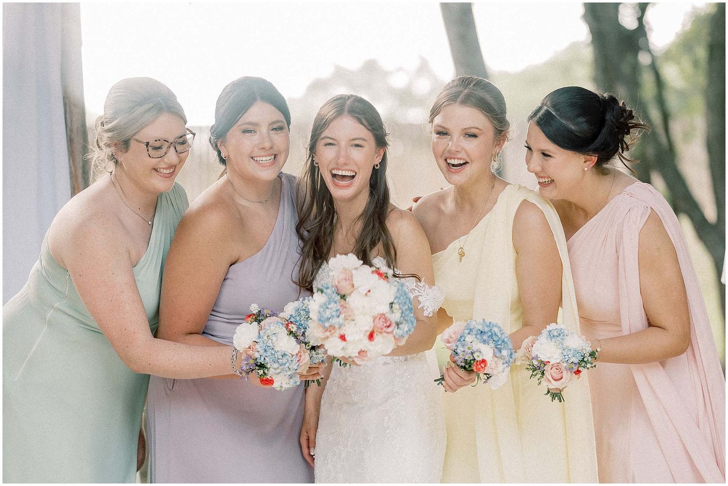 Bridesmaids in pastel pink and lavender dresses share joyful laughter during outdoor wedding photography session.