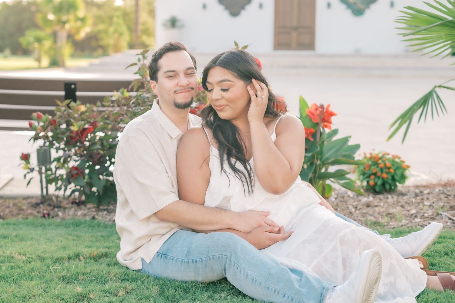 A romantic couple in white outfits sits together on grass near colorful flowers and palm trees in a garden setting.