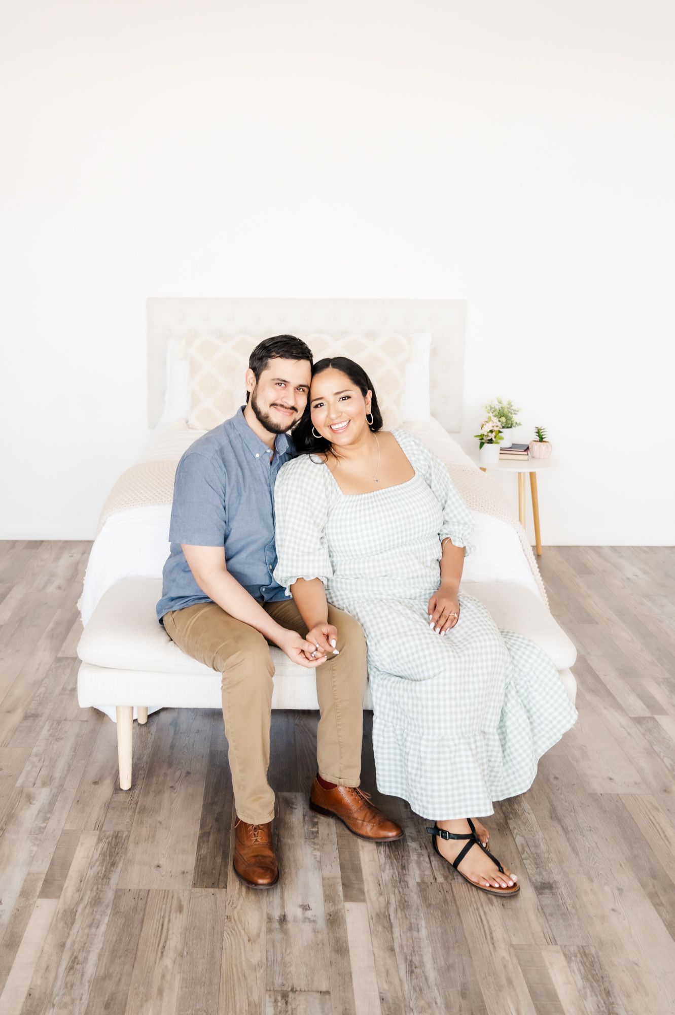 Couple sitting together on white bench wearing blue shirt and checkered dress in bright minimal studio setting.