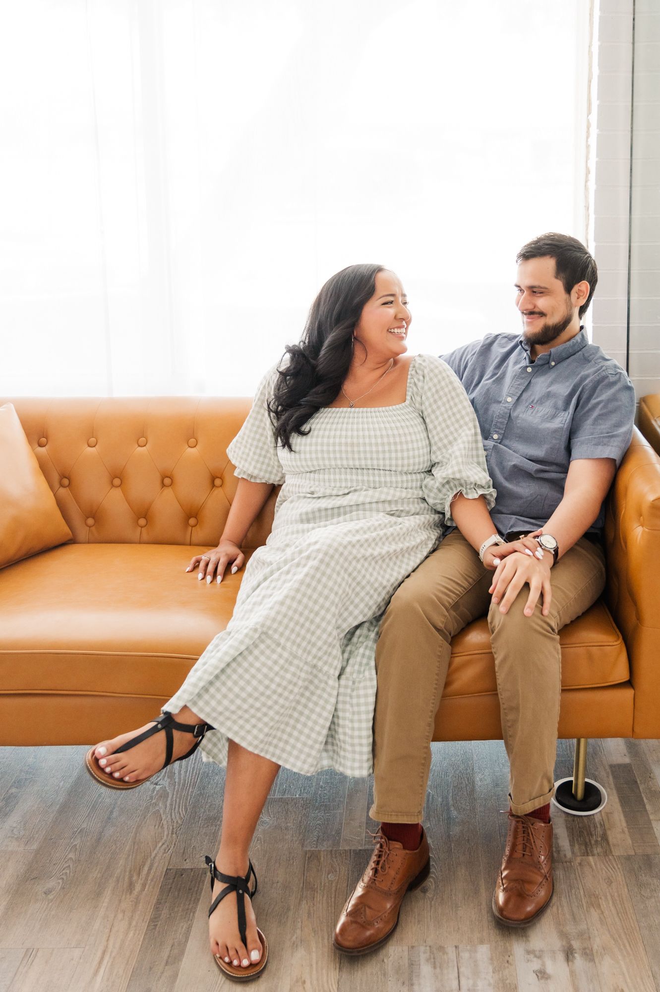 A couple sits together on a tan leather couch, sharing a joyful moment in a bright indoor setting.