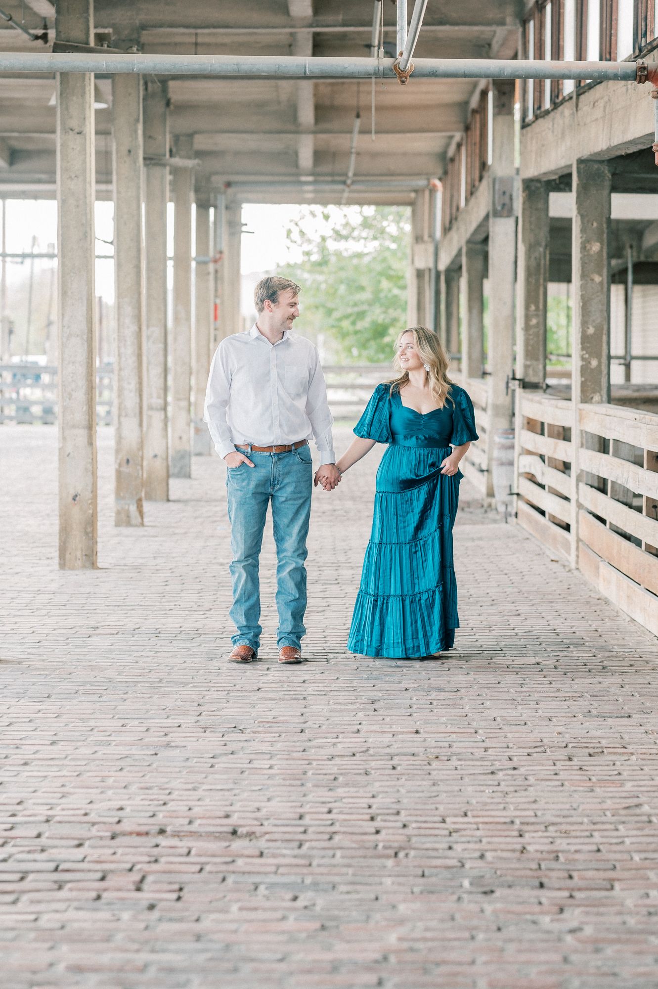 A couple walks together in an industrial space with brick floors, the woman wearing a flowing teal dress.