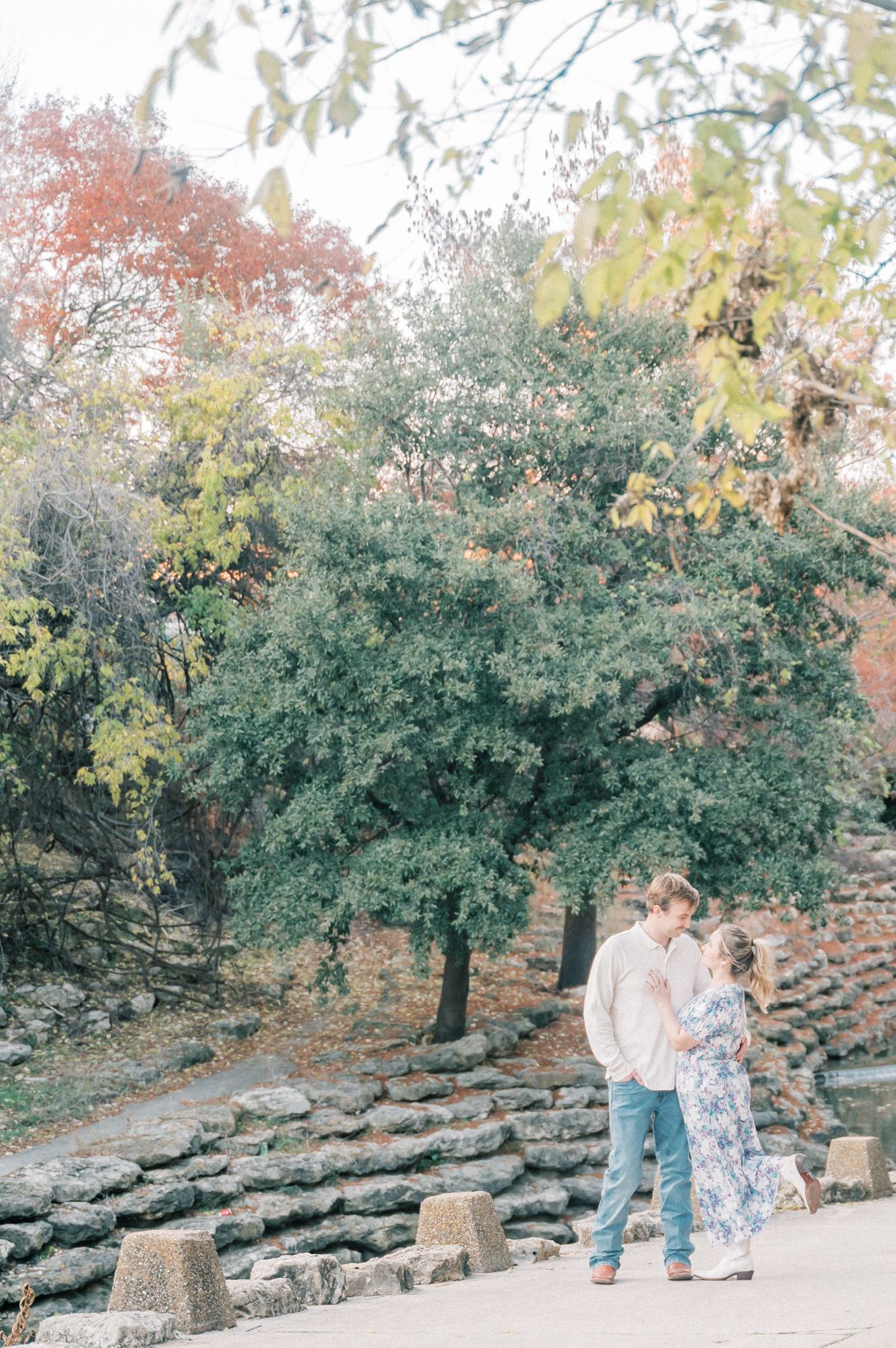 A couple shares a tender moment on a stone path surrounded by lush trees and autumn foliage in a park setting.