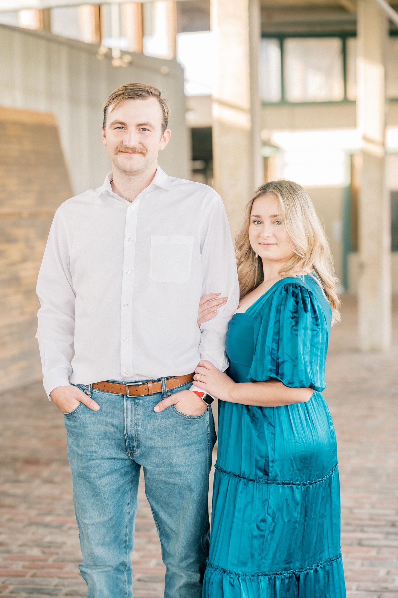 A couple in coordinated white and turquoise outfits pose together for engagement photos in an outdoor setting.