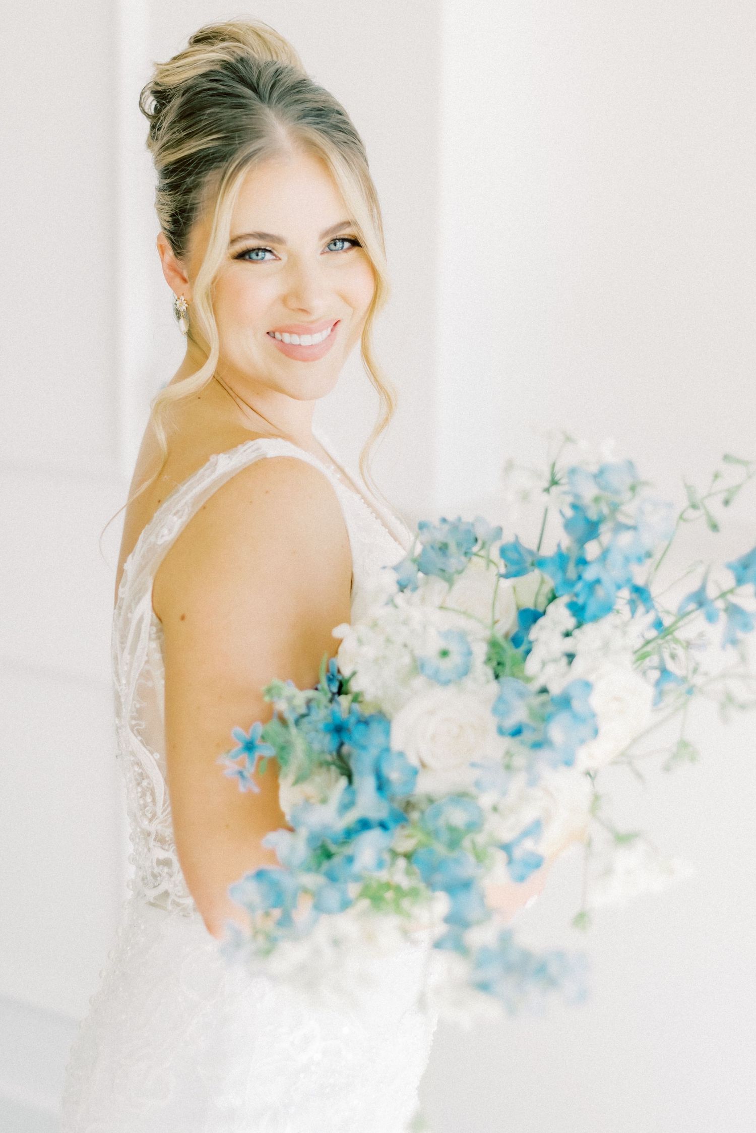 A bride in a lace gown poses with a delicate blue floral bouquet against a bright white background.