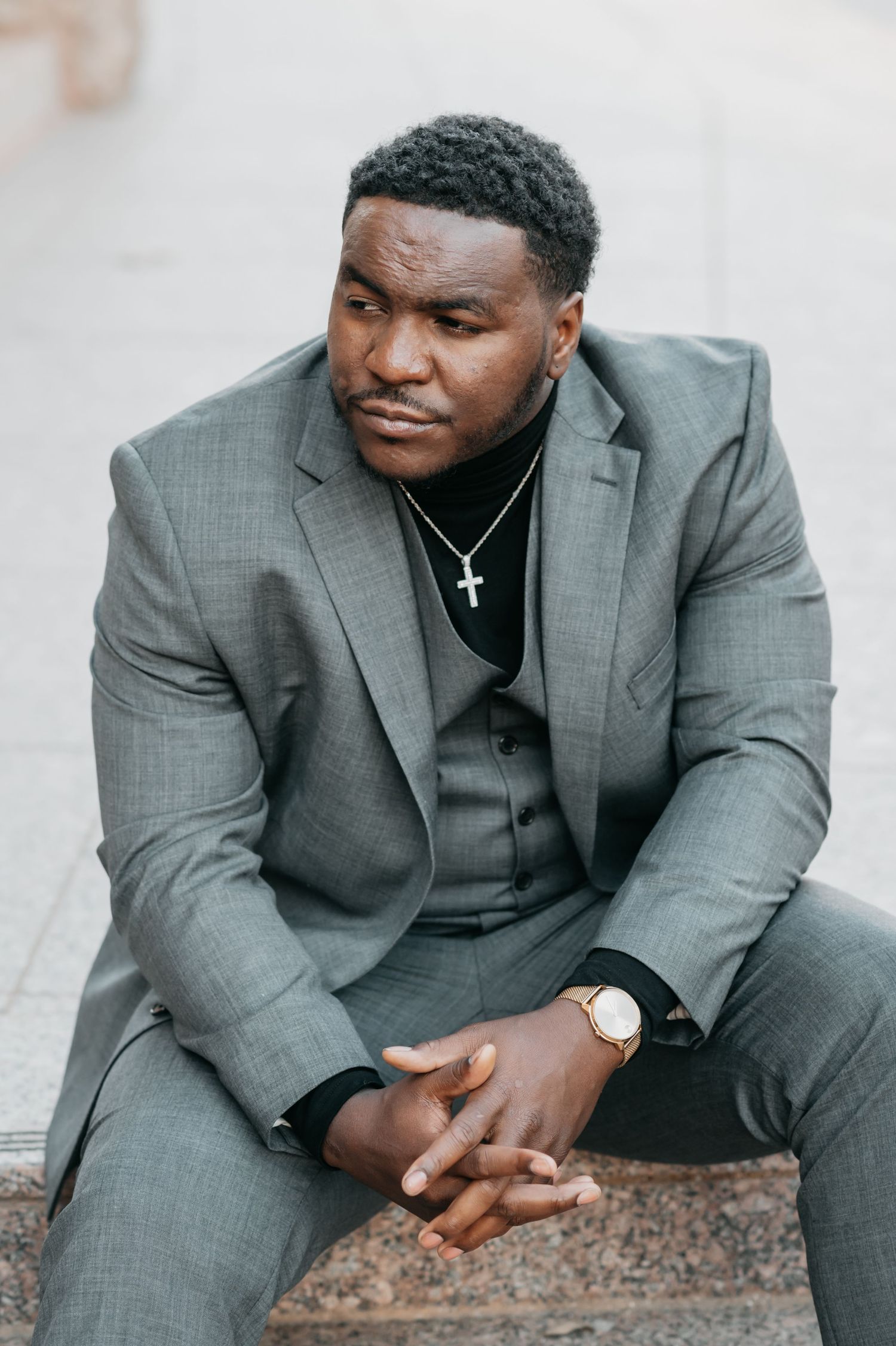 Well-dressed man in gray suit with cross necklace and watch sits thoughtfully on stone steps outdoors.