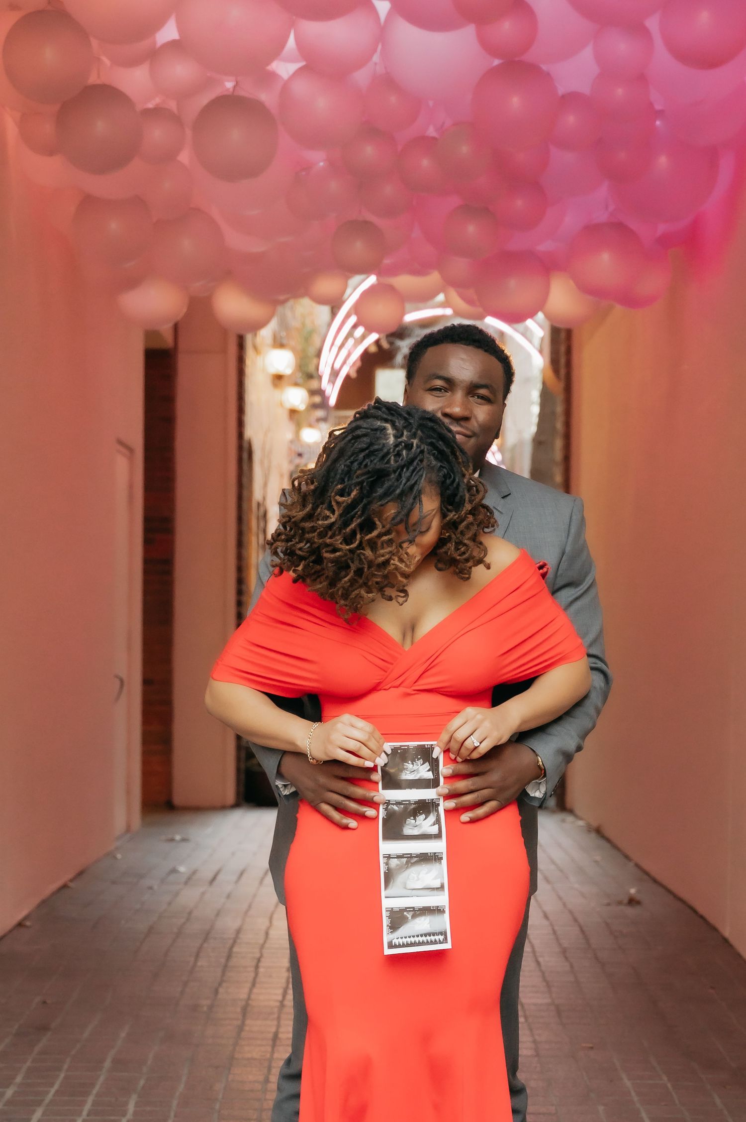 Couple in matching gray suit and red gown holding ultrasound photos under pink balloons, announcing pregnancy.