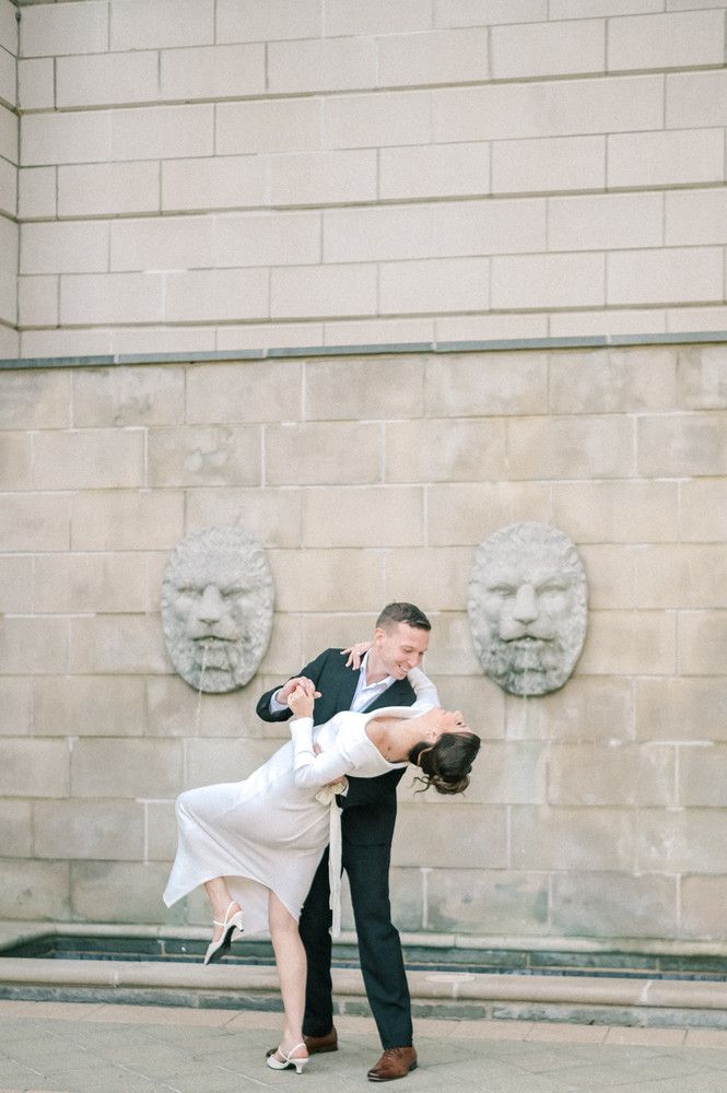 Couple dancing outdoors, man dipping woman in white dress near stone wall with lion sculptures.