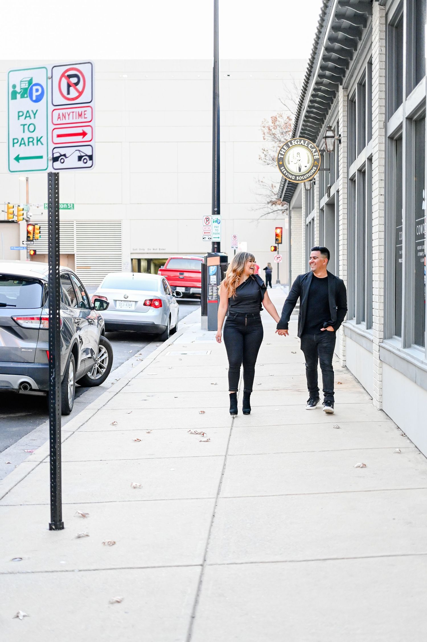 Couple dressed in black holding hands while walking along a sunny downtown sidewalk past a clock tower.