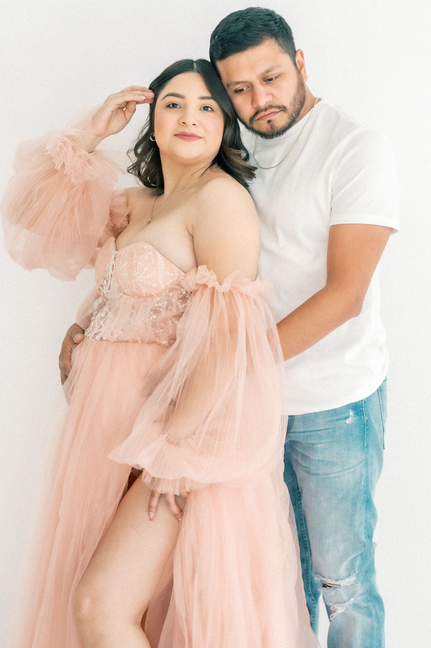 Expecting couple poses in studio, woman in blush tulle gown with partner behind her.
