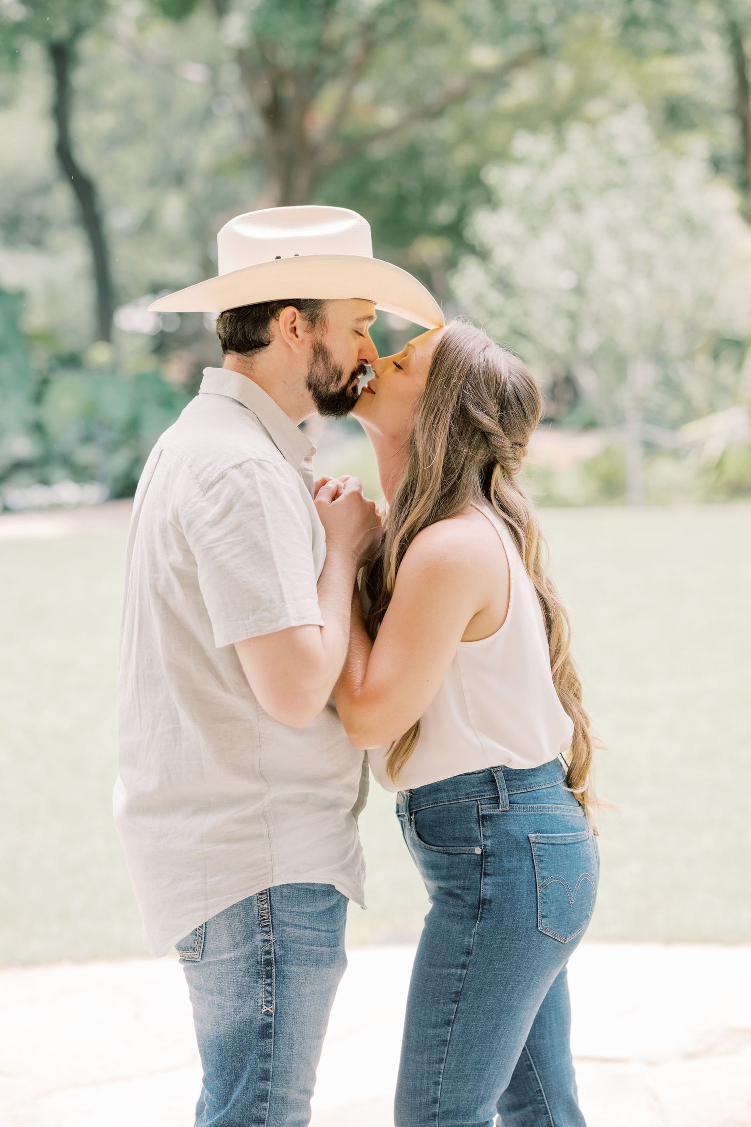 An engaged couple shares a romantic kiss outdoors, man in cowboy hat holding hands with woman in white tank top.