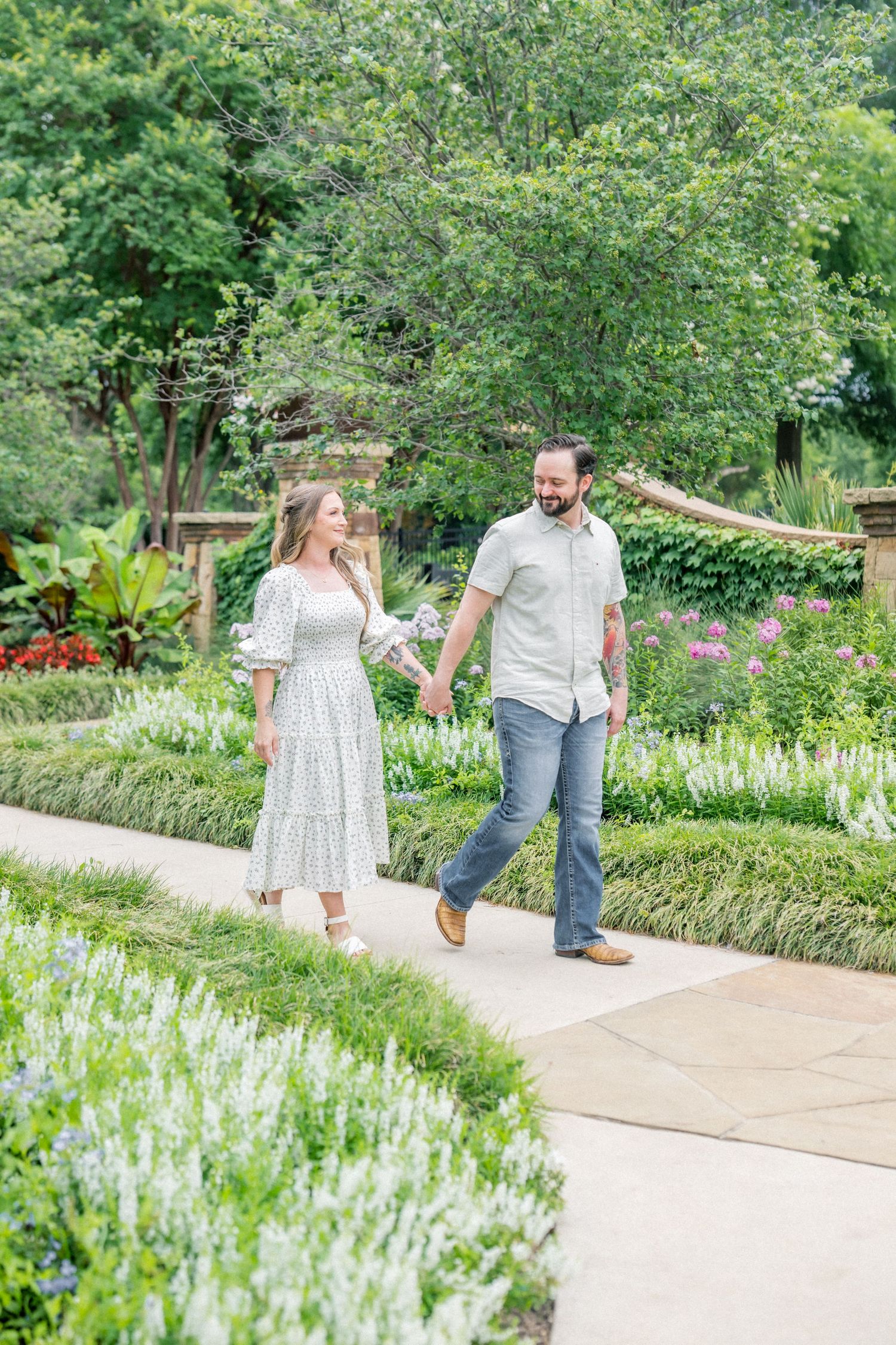 A couple holds hands and walks along a garden path surrounded by lush greenery and colorful flowers on a sunny day.