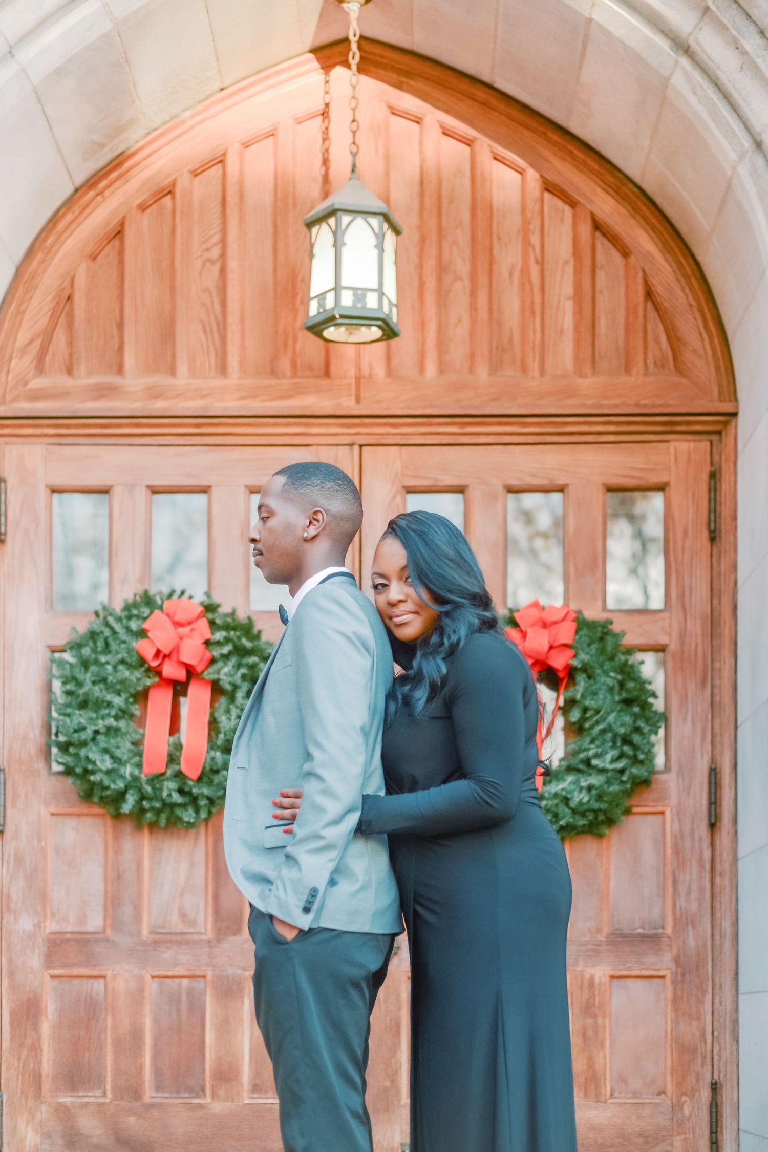 Couple posing in front of wooden church doors adorned with Christmas wreaths and red bows.