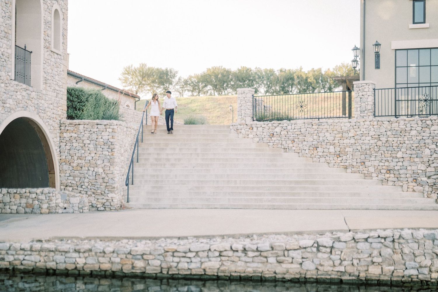 Couple walking up stone steps at an elegant outdoor venue with limestone walls and lush trees in the background.