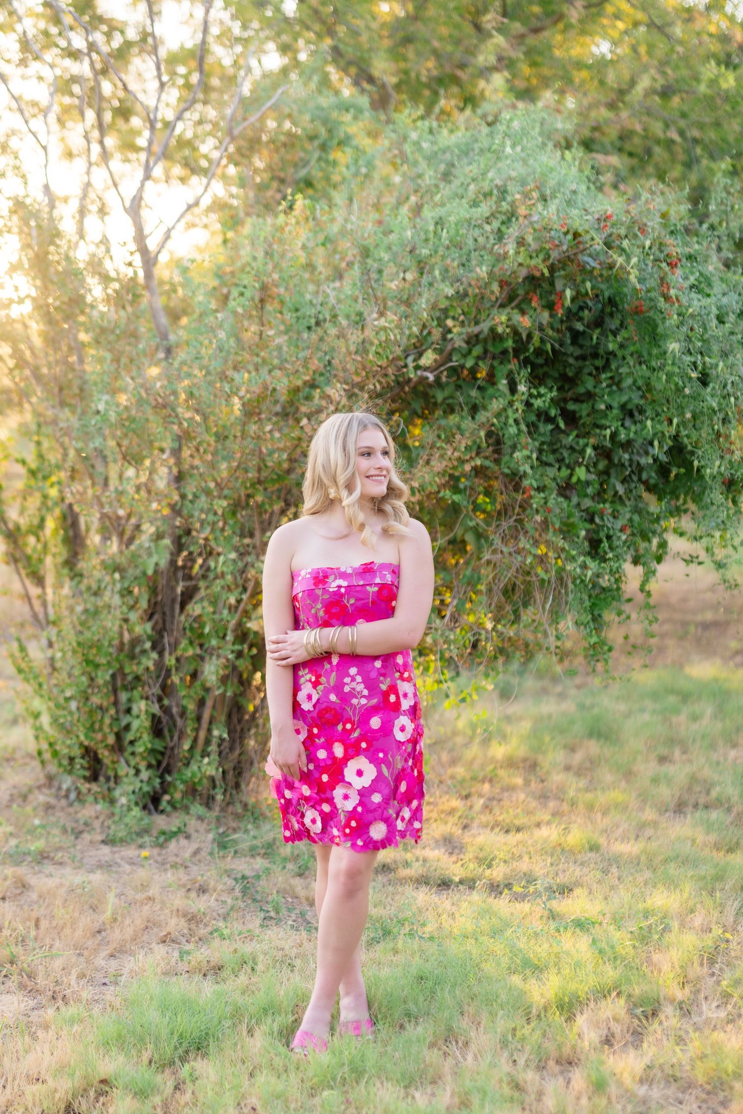 Young blonde woman in pink floral strapless dress standing outdoors in golden sunlight near green trees.