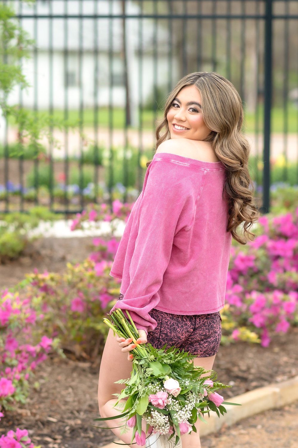 Young woman in pink sweater and floral shorts holding a bouquet of flowers in a garden with pink blooms.