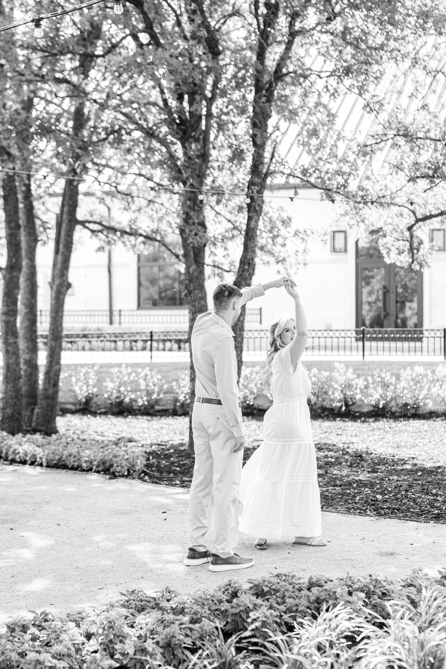 Black and white photo of a couple dancing outdoors under trees in a garden pathway.