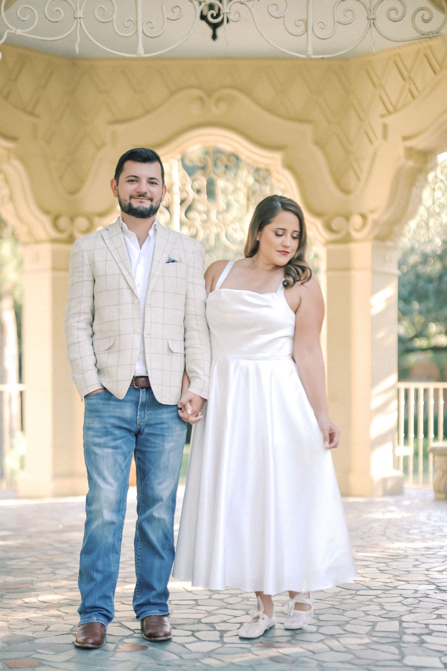 Smiling couple hold hands under an ornate yellow pavilion, she in a white dress and he in a plaid blazer and jeans.