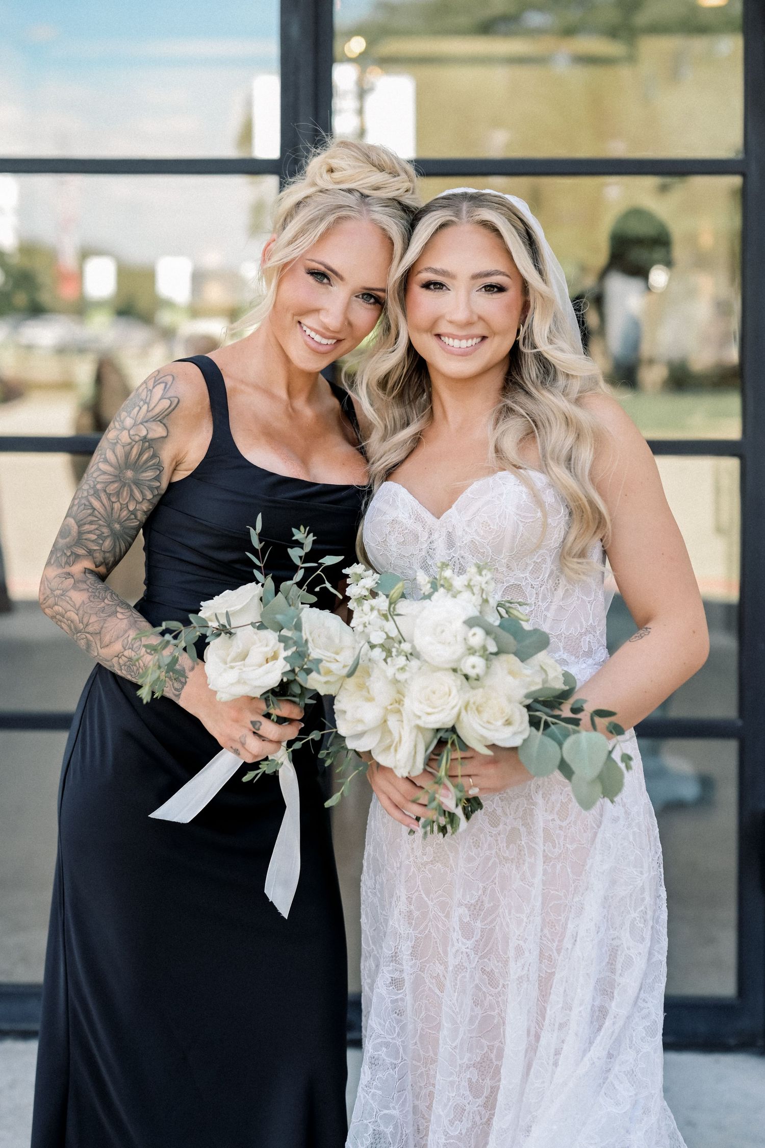 Bride in lace wedding gown poses with tattooed bridesmaid in black dress outdoors.