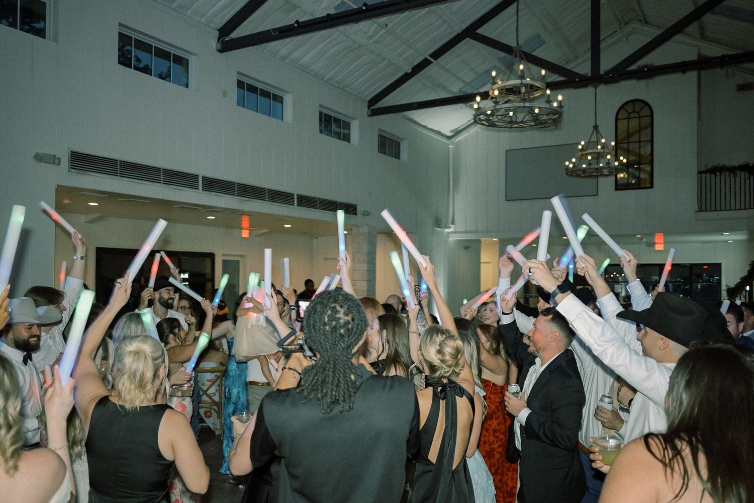 Wedding reception guests dance and wave colorful glow sticks in elegant white venue ballroom.