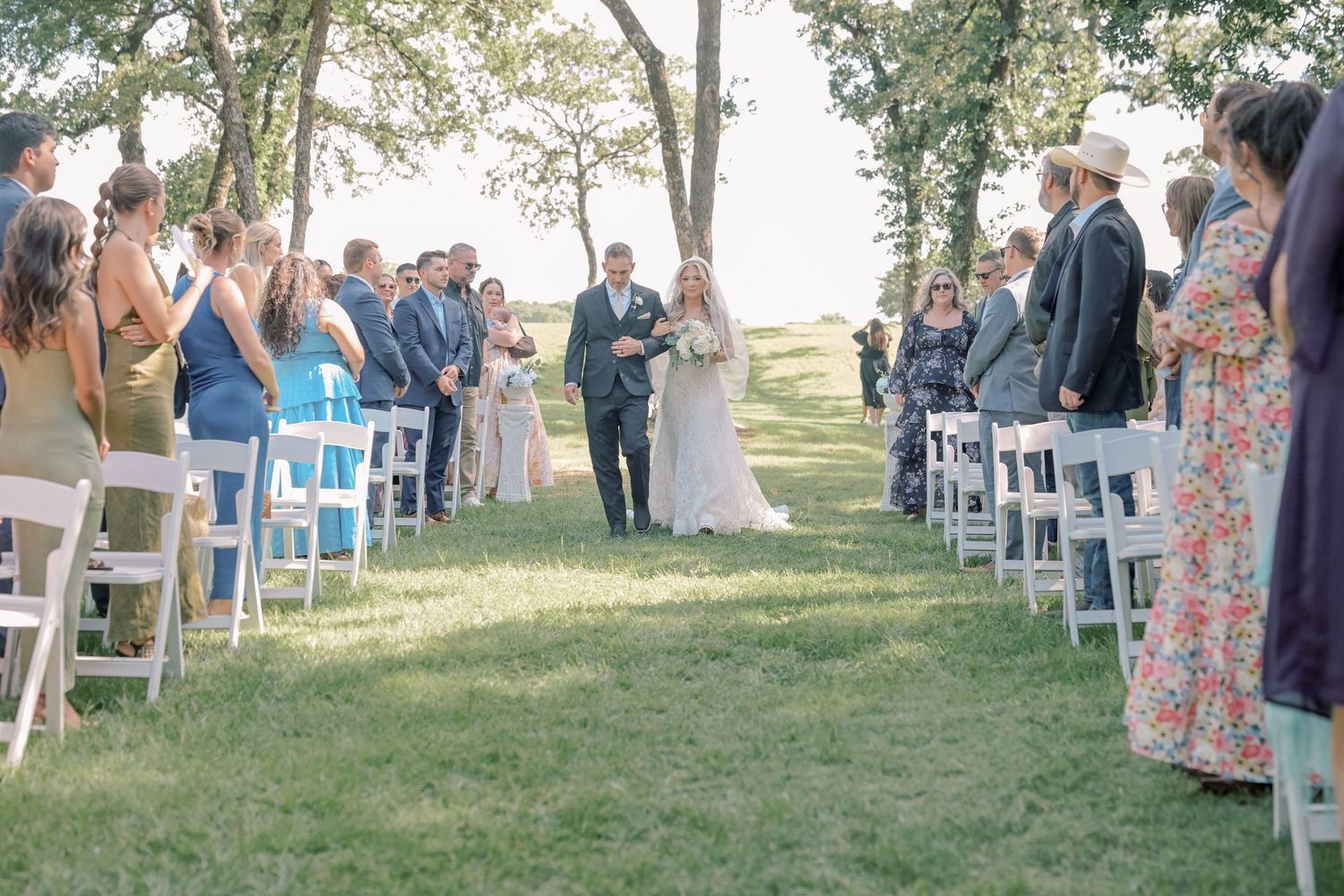 Bride and groom walk down outdoor aisle lined with white chairs and guests during wedding ceremony.
