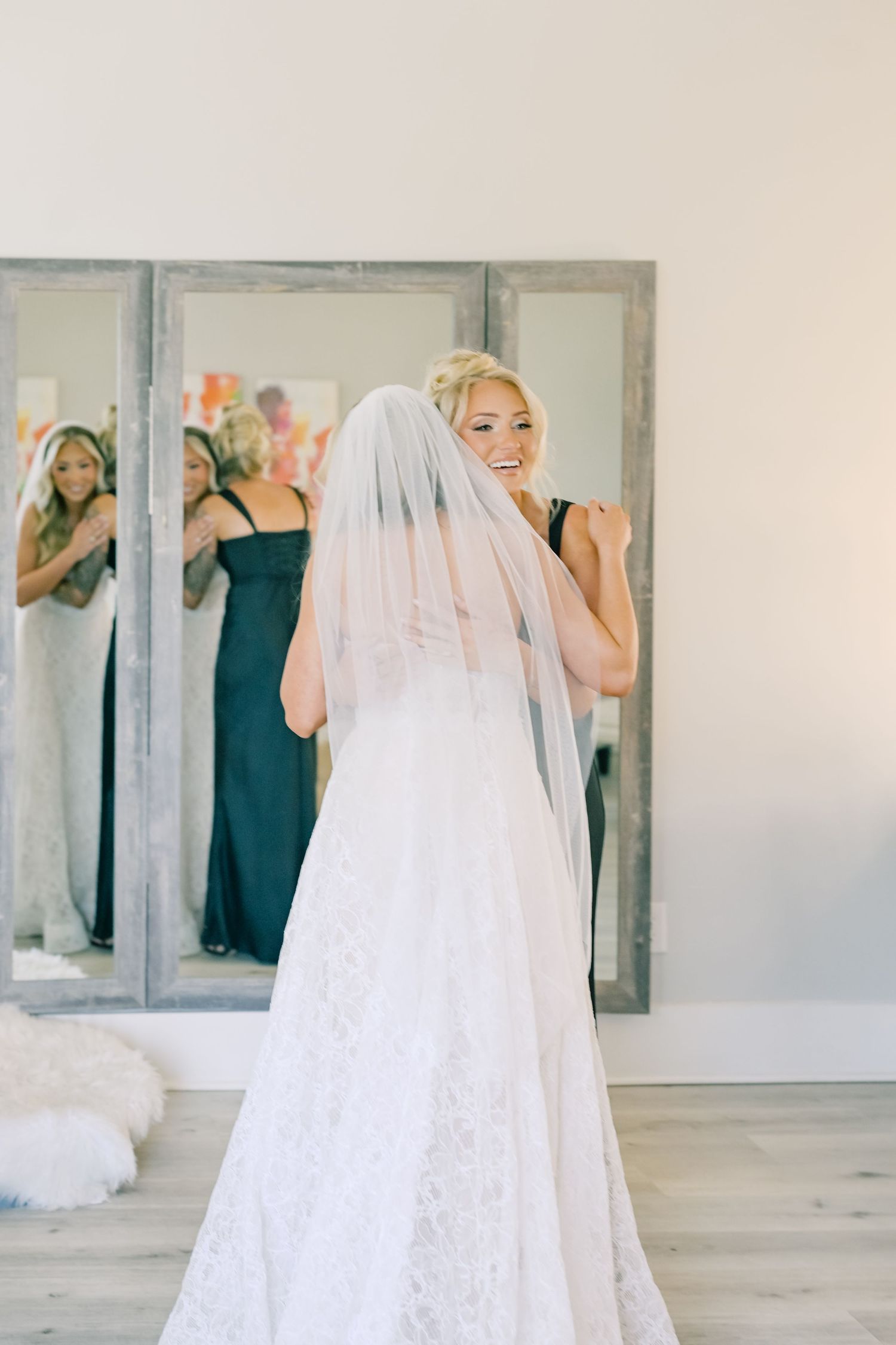 Smiling bride in lace wedding gown and veil poses in front of mirror with bridesmaids in black dresses.