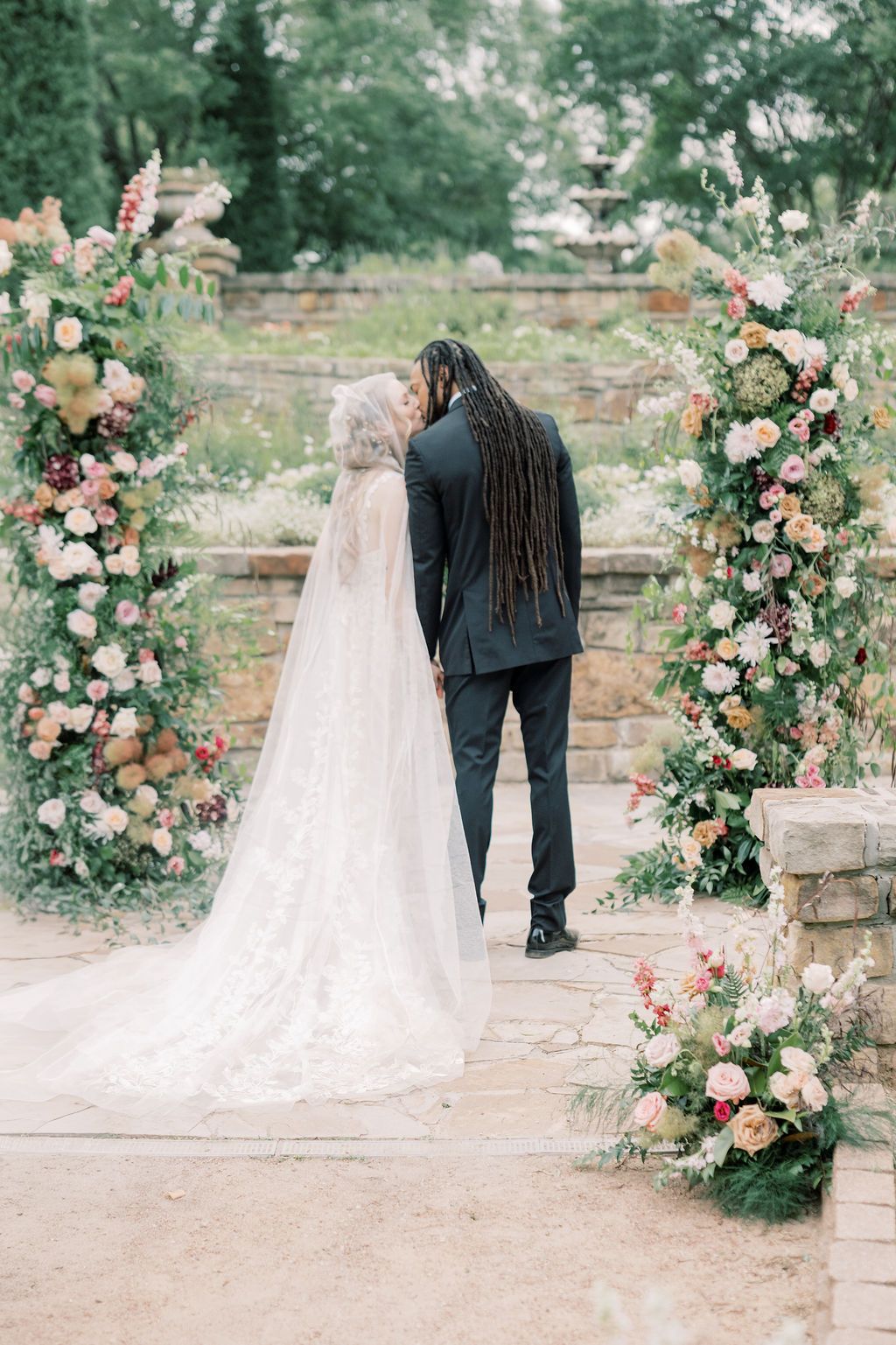 A bride and groom share a kiss at a floral arch altar adorned with pink blooms during an outdoor wedding ceremony.